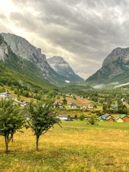 Man sieht ein Tal am Balkan. Im Hintergrund sieht man mehrere Berge, sowie ein kleines Dorf. Im Vordergrund stehen zwei Bäume.