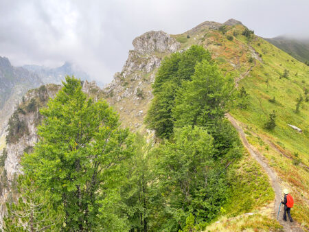 Man sieht einen Berggipfel hinter Wolken verhangen im Bergland des Balkans. Eine Person auf Wanderreise ist klein am Rande des Bildes zu erkennen, während sie die traumhafte Aussicht über Albanien, Nord-Mazedonien oder den Kosovo genießt.