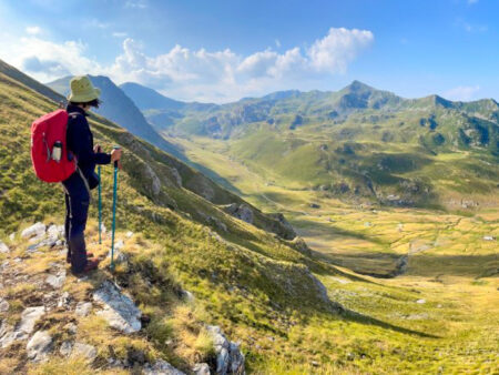 Eine Person auf Wanderreise steht mit ihrem großen, roten Rucksack auf einer Felsvrosprung am Balkan. Sie schaut hinaus auf ein Tal im Herbst oder Frühjahr, und stützt sich auf ihre Wanderstöcke.