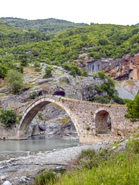 Die Ura e Kadiut, eine bekannte osmanische Steinbogenbrücke im Süden von Albanien ist am Bild zu sehen, wie sie einen Fluss überspannt. Im Hintergrund sieht man das albanische Bergland.