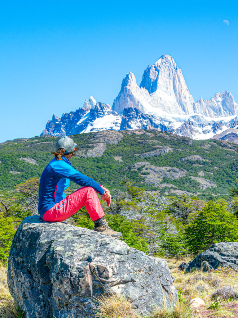 Eine Frau auf Wanderreise sitzt auf einem Felsen und schaut über die Landschaft von Patagonien hinaus. Im Hintergrund ragen die Gipfel des Fitz Roy Bergmassivs auf.