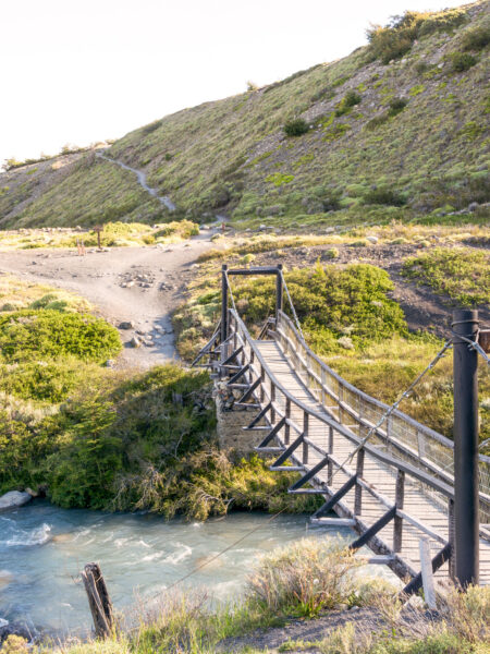 Eine Brücke führt über einen schmalen Fluss in Patagonien. Davor und dahinter führt ein Wanderweg durch die Landschaft.
