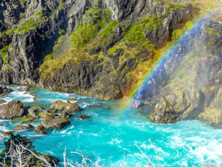 Das Bild zeigt einen Fluss in Patagonien. Das Wasser scheint türkis in die Kamera und ein kleiner Regenbogen ist über dem Wasser zu erkennen, was auf einen Wasserfall außerhalb des Bildes schließen lässt.