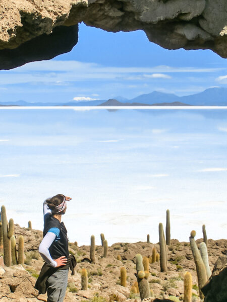 Eine Frau auf Wanderreise steht zwischen großen Kakteen und schaut durch ein Loch in einer Felsformation hinaus auf den Salar de Uyuni, den größten Salzsee der Welt, der sich von Bolivien bis nach Peru erstreckt.