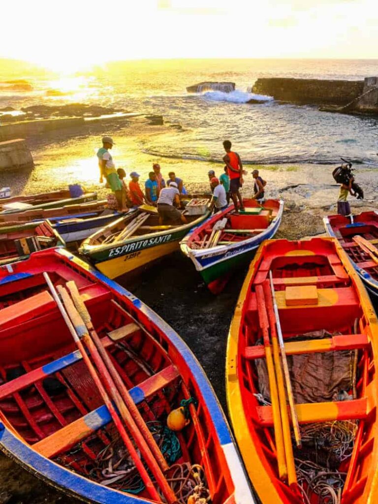 Mehrere Boote liegen vor der Küste einer Insel von Kap Verde. Sie sind beschienen vom Licht der untergehenden Sonne und mehrere Personen sitzen um ein Boot herum.