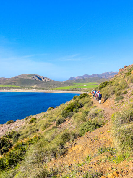 Man sieht die Küste Südspaniens. Auf einem schmalen Wanderweg durch die karge Landschaft gehen mehrere Personen auf Wanderreise.