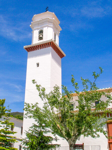 Der Turm der Kirche Iglesia Parroquial de San Roque ragt in den kräftigen blauen Himmel über der Region Las Alpajurras in Spanien auf. Im Vordergrund kann man mehrere Bäume sehen.