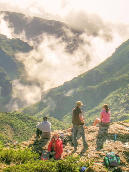 Vier Personen auf Wanderreise stehen bzw. sitzen auf an einem Felsvorsprung entlang einer Wanderroute auf der Atlantik-Insel La Gomera. Im grünen Tal unter ihnen hängen Wolken und Nebel, als sie sich die erste Paus auf ihrer Wanderung des Tages gönnen.