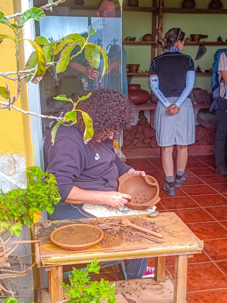 Eine Einheimische auf La Gomera sitzt vor einem Geschäft und mahlt Gewürze in einer Schale aus Ton. Im Hintergrund sieht man eine Touristin auf Wanderreise auf der Atlantik-Insel.