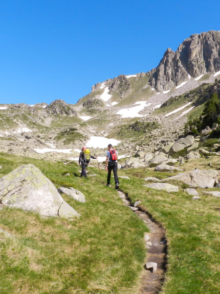 Wir sehen zwei Wanderreisenden auf einem Wanderweg durch die Pyrenönen in Nordspanien. Im Hintergrund sieht man die Berge, dich zu Teil noch von Schnee bedeckt sind. Beide Personen tragen Wanderausrüstung.