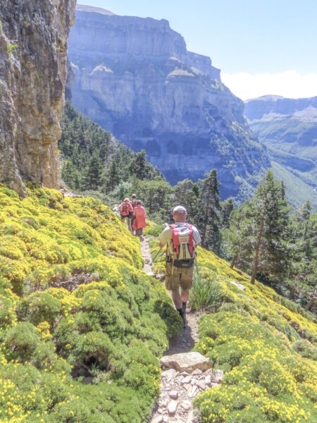 Zwei Personen auf einer Wanderreise folgen einem schmalen Wanderweg entlang eines Abhangs. Im Hintergrund erkennt man den Nationalpark Ordesa y Monte Perdido in den Pyrenäen Nordspaniens.