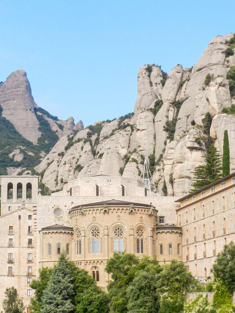 Das Bild zeigt das Kloster Montserrat, das hoch in den Felsen der Pyrenäen thront. Hinter der Fassade des Gebäudes ragen die malerischen Berge Nordspaniens auf.