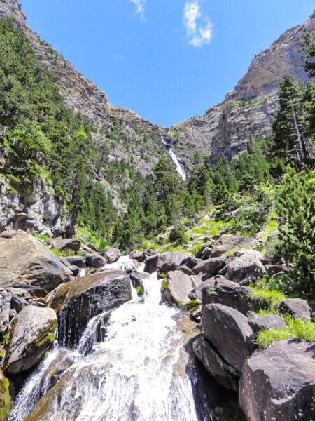 Wir sehen einen reißenden Bach, der sich über kleine Felsen in seinem Bett durch den Nationpark Ordesa in Nordspanien schlängelt. Im Hintergrund ragen Felswände auf.