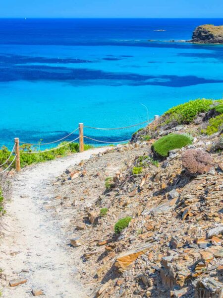 Auf dem Foto sieht man einen Wanderweg auf der Mittelmeerinsel Menorca. Dahinter breitet sich das blaue Meer aus, das man von der Anhöhe auf dem Weg aus sehen kann.
