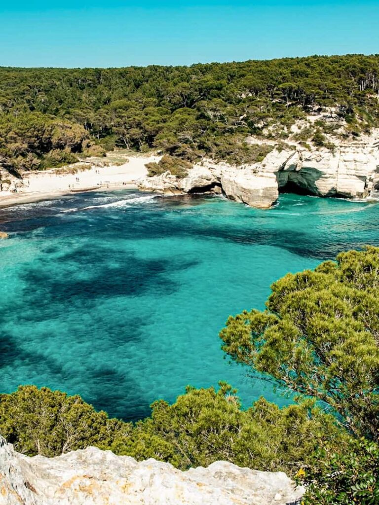 Die Cala Mitjana auf Menorca besticht durch ihr türkisblaues Wasser. Mehrere Büsche im Vordergrund verstecken sie vor Blicken und bieten den Wandergruppen auf ihrer Reise einen abgeschiedenen Ort zum Baden, abseits des Massentourismus.