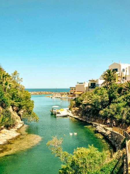Ein Fluss auf Menorca windet sich durch das Bild. Ein Haus steht im Hintergrund und mehrere Büsche im Vordergrund.
