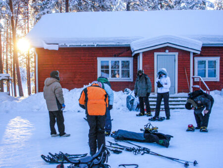 Eine kleine Gruppe, die zum Schneeschuhwandern nach Finnland gekommen ist, bekommt vor einem roten Haus in der finnischen Taige ihre gestellte Ausrüstungen erklärt und beigebracht.