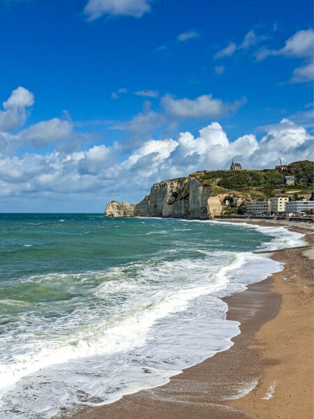 Auf dem Landschaftsfoto sieht man die berühmten Kreidefelsen von Étretat an der Alabasterküste in der Normandie. Im Vordergrund brechen die Wellen am Strand.