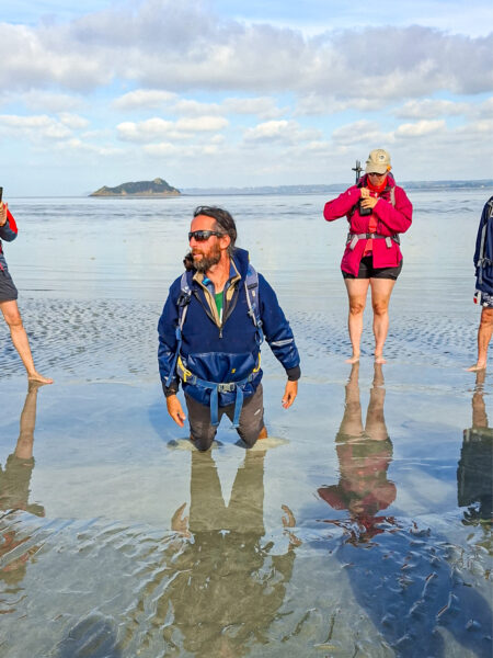 Eine kleine Gruppe auf einer Wanderreise durch die Bretagne steht um ihren Wanderführer herum, der bis zu den Knien im Watt vor Mont-Saint-Michel feststeckt.