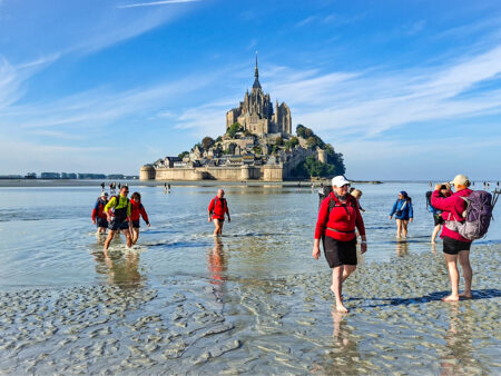 Eine kleine Gruppe auf einer Wanderreise in der Bretagne geht auf dem Bild durch das Watt vor der französischen Nordküste. Im Hintergrund sieht man die Ortschaft Mont-Saint-Michel.