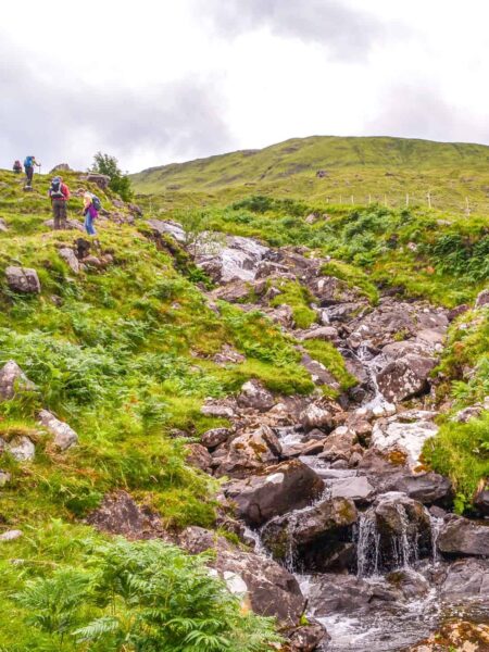 Mehrere Personen auf Wanderreise gehen einen Hang neben einem kleinen Bach in Connemara, Irland hinauf.