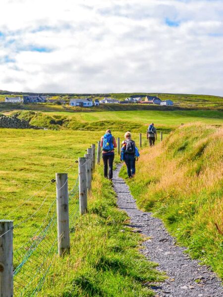 Drei Personen folgen einem irischen Wanderweg in Richtung einer kleinen Ortschaft.