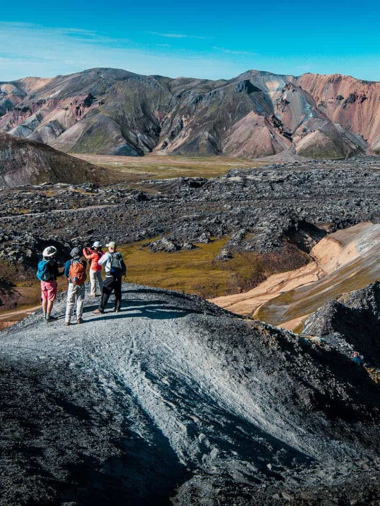 Eine kleine Gruppe auf einer Wanderreise durch Island steht auf einem schwarzen Sandhügel inmitten der bunten Berge der in der vulkanischen Landschaft von Landmannalaugar im isländischen Hochland.