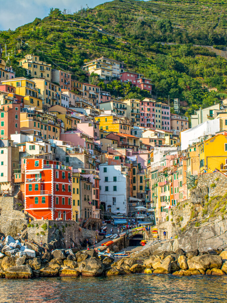Ein vom Wasser aufgenommenes Foto der Altstadt von Riomaggiere, einem der Cinque Terre in Ligurien. Die Stadt besticht durch bunte Häuser, die an einem Hang in Ligurien errichtet sind.