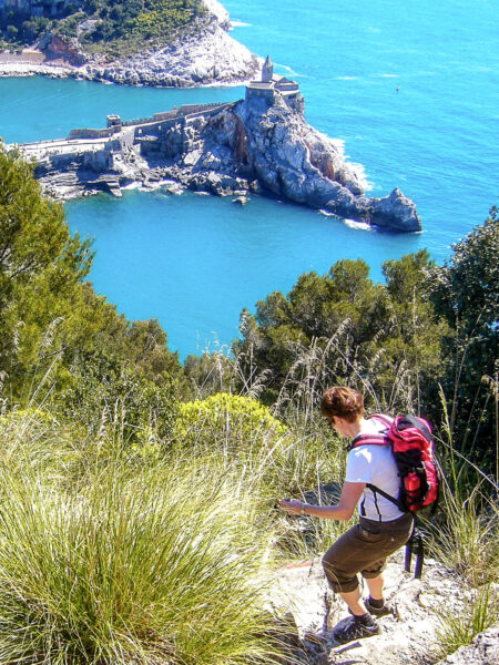 Ein Frau auf Wanderreise durch Ligurien geht einen steilen Wanderweg hinab. Im Hintergrund sieht man die Küste, sowie einen Teil von Portovenere.