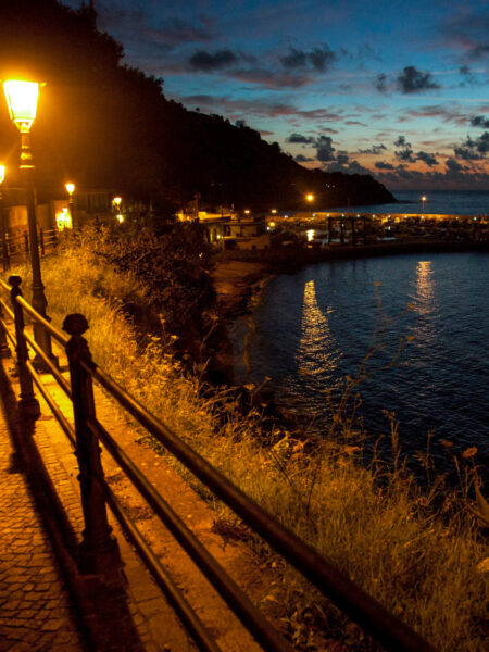 Ein Steg bei Nacht in Italien. Mehrere Laternen leuchten an dem Geländer, das Menschen auf Wanderreise vom Meer trennt.