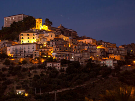 Man sieht die Ortschaft Castellabate in der Bucht von Salerno in Italien bei Nacht. Die Stadt ist warm beleuchtet und ragt auf einem Hügel über die Landschaft.