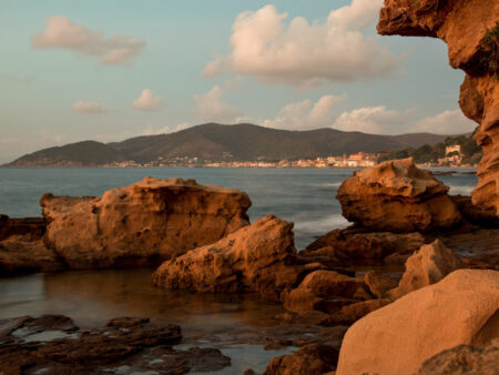 Man sieht mehrere Felsen im Meer liegen. Dahinter lässt sich eine kleine, italienische Ortschaft an der Küste einer Bucht erkennen.