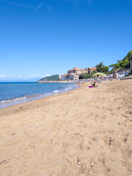 Man sieht einen Sandstrand im Cilento. In der Distanz liegen wenige Leute unter Schirmen und man kann auch eine kleine Ortschaft des Cilento im Hintergrund erkennen.