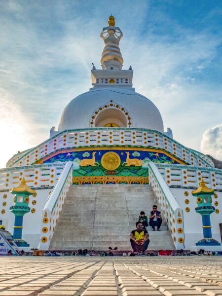 Das Bild zeigt die Shanti Stupa in Ladakh im Norden von Indien. Auf der Stufen der weißen Stupa sitzen drei Einheimische aus Nordindien.