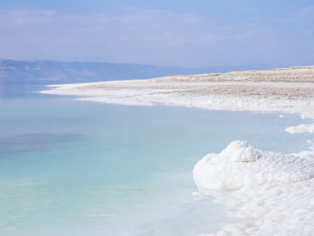 Ein Foto des Toten Meers in Jordanien. Man sieht die Küste des stark salzhaltigen Gewässers, wo sich auch kleine Haufen aus Salz auftürmen.