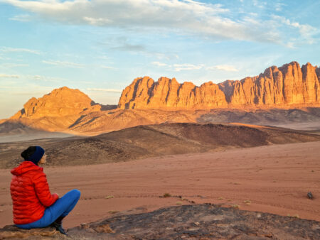 Eine Frau auf einer Wanderreise mit Weltweitwandern, sitzt in der Wüste von Jordanien. Sie befindet sich im Wadi Rum, im Hintergrund sieht hoch in den Himmel ragende Felswände.