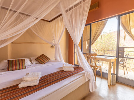 Zimmer der nachhaltig betriebenen Unterkunft Feynan Ecolodge mit einem gemütlichen Bett mit Kissen, Moskitonetzen, einem kleinen Schreibtisch mit Stuhl und großen Fenster zum Balkon mit Blick in Wadi Feynan.