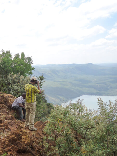 Ein Wander-Reisender und ein lokaler Guide stehen an einer Böschung am Hannington-Hike und schauen hinunter auf einen See in Kenia.