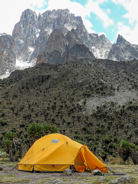 Ein Zelt von Wanderreisenden steht vor dem Hintergrund des Mount Kenya. Es ist gelb und hebt sich klar von dem Hintergrund der schartigen Berglandschaft ab.