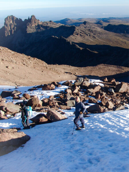 Zwei Wanderreisende gehen den Weg vom Gipfel des Mount Kenya hinunter. Sie hinterlassen Spuren im Schnee auf dem zweithöchsten Berg in Afrika. Hinter ihnen kann man große Landschaftsabschnitte in Kenia erkennen.