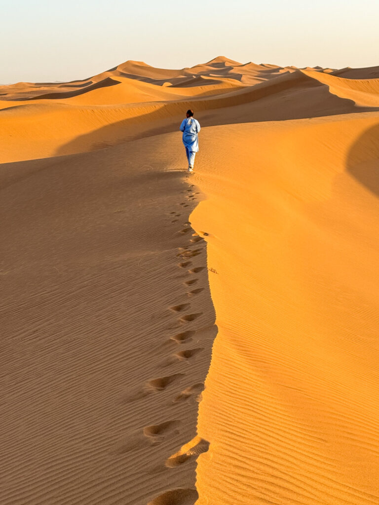 Einen Person auf Wanderreise geht am Grat einer Sanddüne in der marokkanischen Wüste entlang. Man sieht sie von hinten und sie hinterlässt Fußspuren im warmen Licht der untergehenden Sonne.