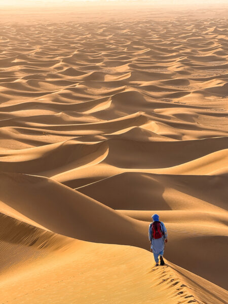Eine Person auf Wanderreise steht auf einer Sanddüne in der Wüste Marokkos. Die Sonne geht gerade auf oder unter und die Person schaut ihr entgegen.