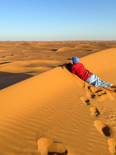 Eine Person auf Wanderreise liegt auf dem höchsten Punkt einer Sanddüne in Marokko. Sie trägt eine rote Jacke von Weltweitwandern sowie einen blauen Turban.