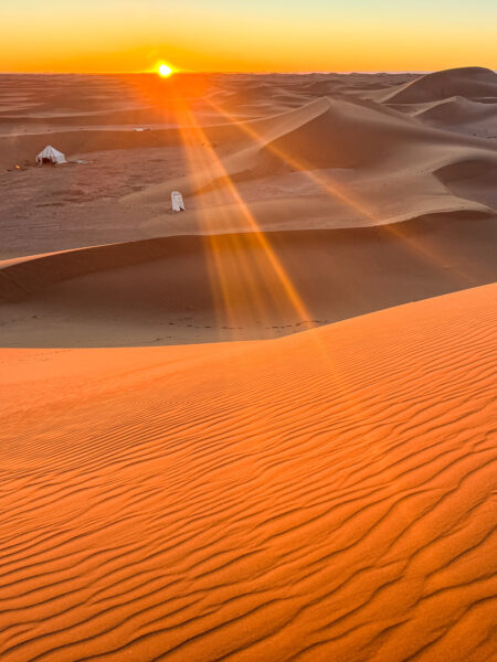 Man sieht die auf- oder untergehenden Sonne in der Sahara. Im Hintergrund sieht man ein Berberzelt zwischen den Sanddünen, das dort für die Reisenden und Guides einer Weltweitwandern-Reise errichtet wurde.