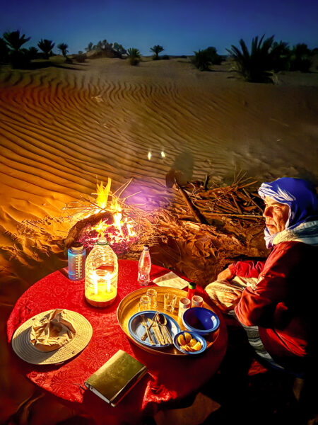 Ein Weltweitwandern-Guide bereitet nach Einbruch der Dämmerung neben einem kleinen Lagerfeuer die Mahlzeit für die Reisegäste vor. Er lächelt freundlich in die Kamera.