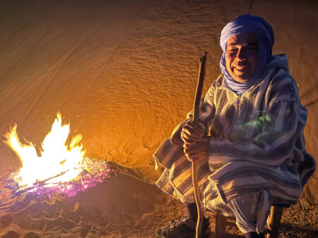 Eine Person in traditioneller marokkanischer Kleidung mit Turban hockt neben einem Lagerfeuer im Camp einer Reise von Weltweitwandern.