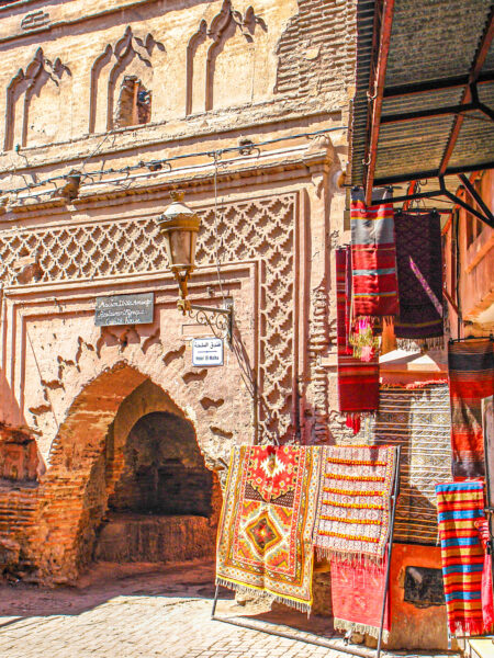 Im Fokus des Bilds steht ein Durchgang eines Gebäudes im Süden von Marokko. Rechts davon hängen einige bunte Teppiche.