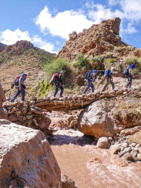 Eine kleine Reisegruppe überquert einen schmalen Bach im Hohen Altas in Marokko. Hinter ihnen kann man mehrere Felsformationen im Gebirge erkennen.
