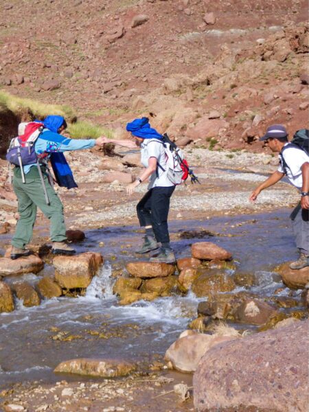 Eine Wanderin auf einer Trekkingreise in Marokko hilft einer anderen Person in ihrer Gruppe dabei, einen Bach im Hohen Atlas auf einigen losen Felsen in dessen Bett zu queren.