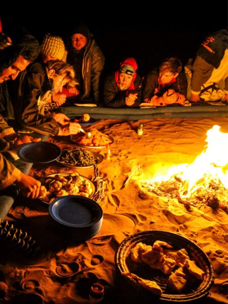 Mehrere Personen auf Wanderreise – Reisende, aber auch Guides – sitzen um ein Lagerfeuer, das im Zentrum eines Trekkingcamps in der Sahara brennt. Die kleine Gruppe und ihre lokalen Guides teilen auf dem Bild Essen und Geschichten.